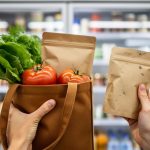 Hands selecting fresh seasonal produce from a reusable tote in a grocery store aisle, with shelves blurred in the background.